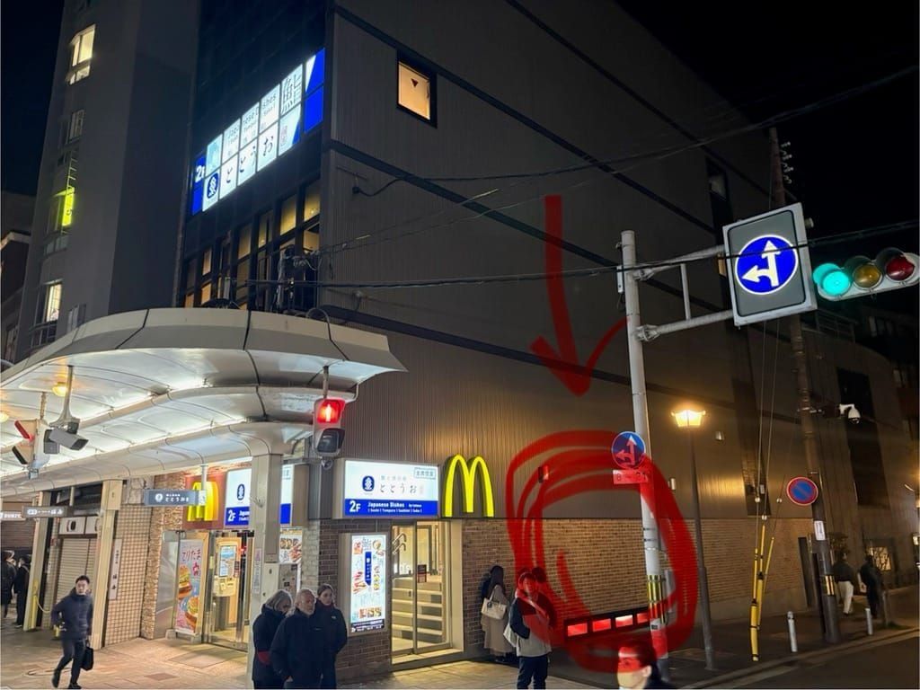 Night view outside Hankyu Kyoto-Kawaramachi Station next to McDonald's, with a red circle marking the sidewalk waiting spot beside the wall.