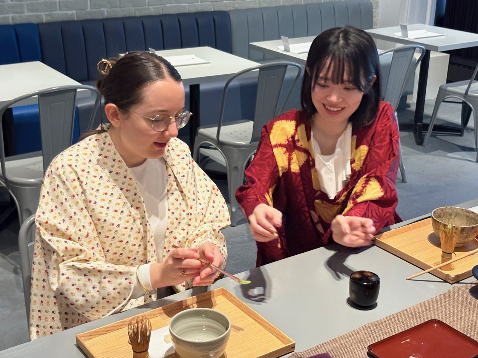 A guest scooping matcha with a bamboo tea scoop, sensei smiling beside her